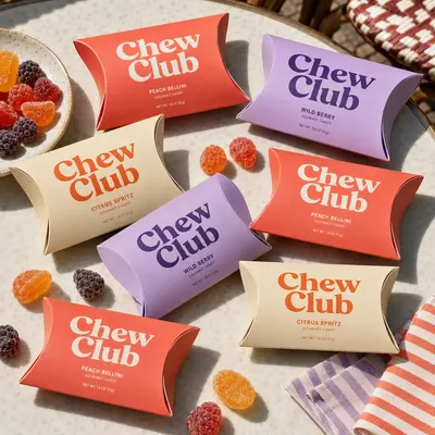 Custom candy pillow boxes in coral, cream, and lavender with "Chew Club" branding, shown grouped closed on a table with loose gummy candies.