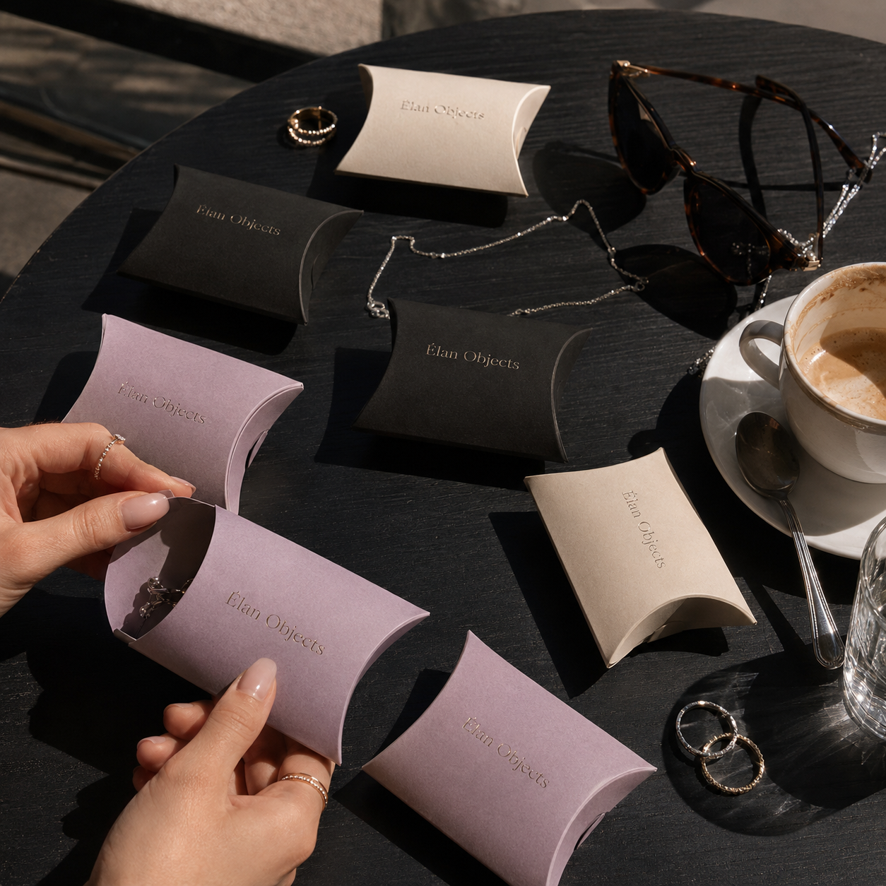 Custom jewelry pillow boxes in cream, black, and lavender with debossed "Élan Objects" branding, shown grouped on a table with rings and a necklace.