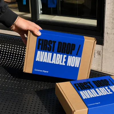 Custom launch box in blue and kraft with "First Drop / Available Now, Common Signal" branding, held and stacked outdoors.