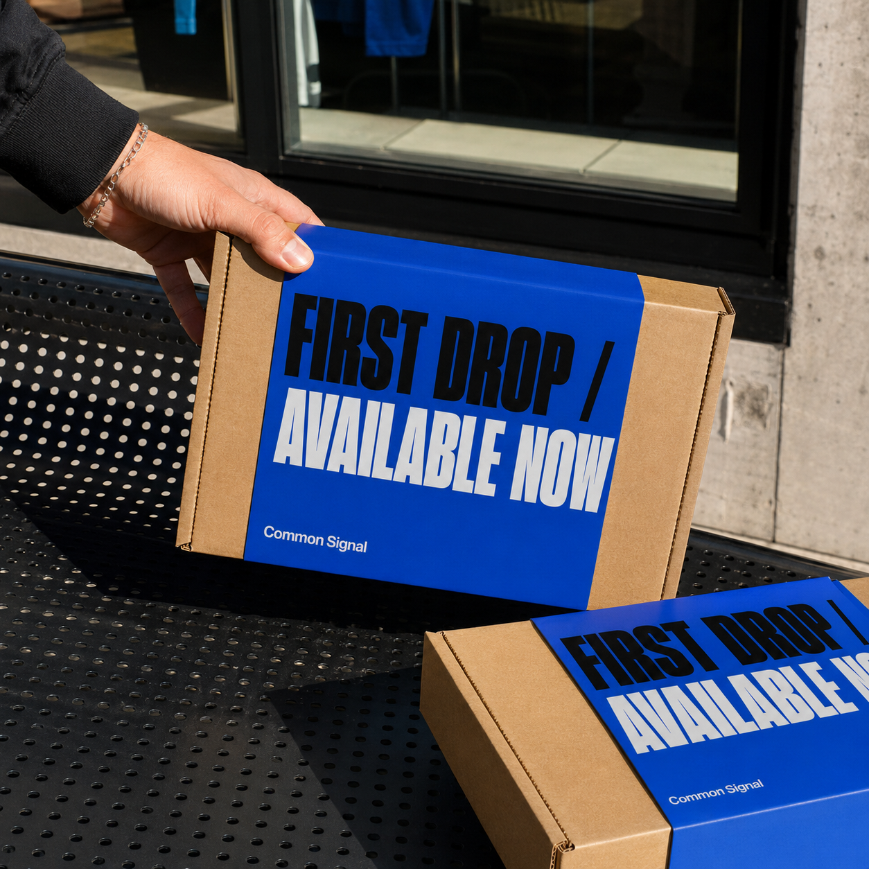 Custom launch box in blue and kraft with "First Drop / Available Now, Common Signal" branding, held and stacked outdoors.