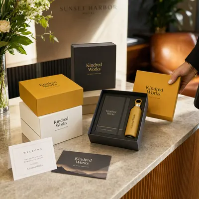 Custom promotional gift boxes in black, gold, and white with "Kindred Works" branding, stacked on a counter with one open showing tissue paper and a notebook inside.