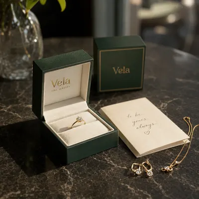 Custom ring box in dark green with gold "Vela" foil branding and cream interior, shown open with a ring inside, closed box and jewelry pieces beside it.