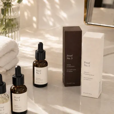 Custom serum box packaging in dark brown and cream with "Proof No. 3" branding, shown upright beside amber dropper bottles.