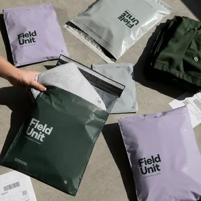 Ecommerce poly mailers in lavender, silver, and dark green, self-seal construction with a tear strip, one opened to reveal a folded garment inside.