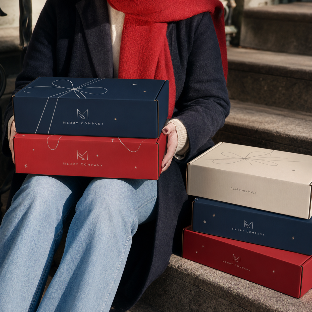 Holiday mailer boxes in navy, red, and gold with a ribbon-bow print, corrugated shipper-box construction, shown stacked and held.