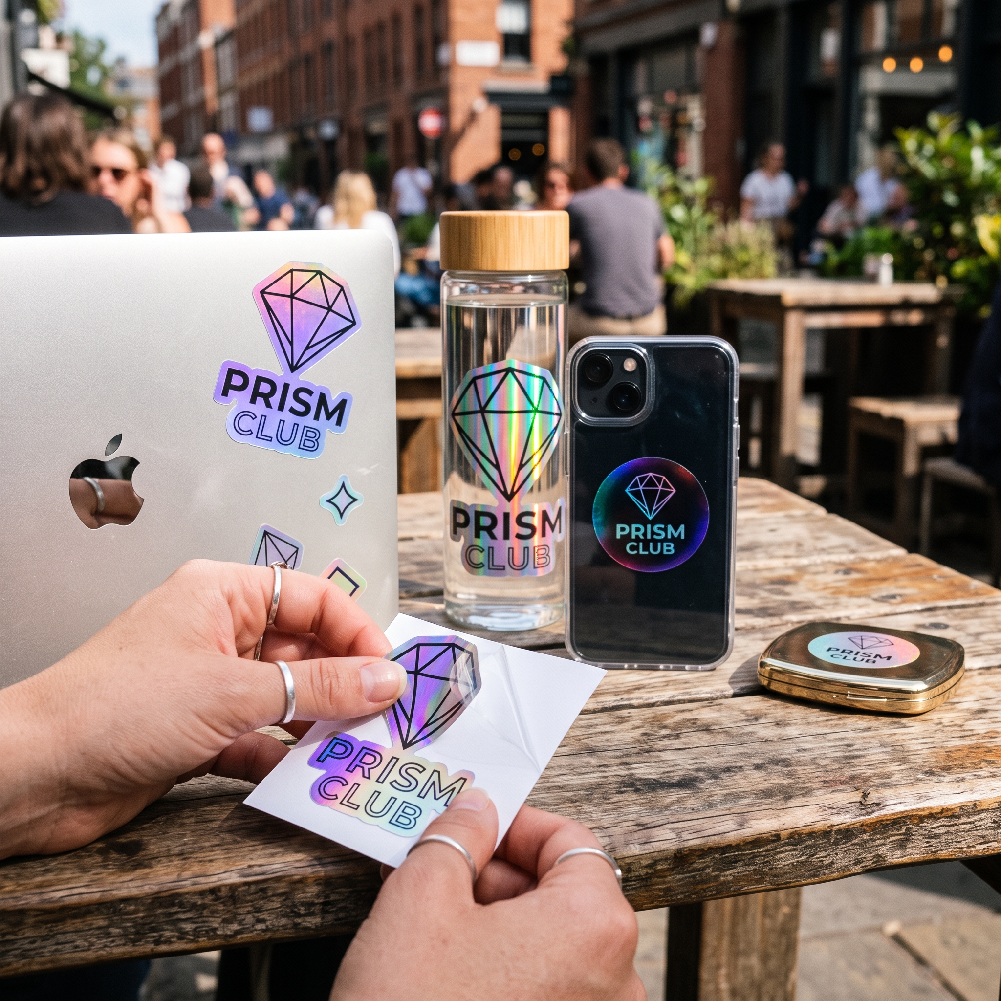 Holographic stickers in diamond and star die-cut shapes with an iridescent finish, applied to a laptop lid, water bottle, phone case, and compact mirror.