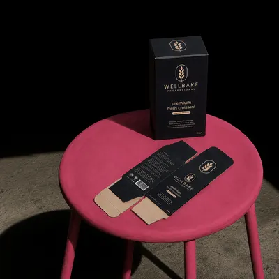Custom reverse tuck box in matte black with gold wheat emblem, shown upright and flattened/unfolded on a pink stool.